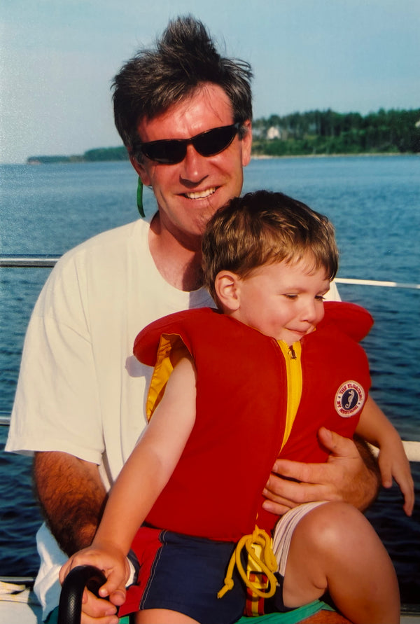 Man holding a child wearing a red life jacket by a body of water.